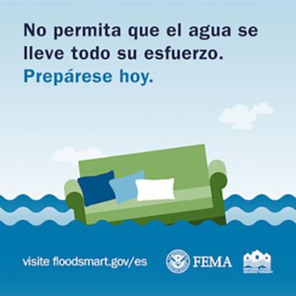 Un sofá verde con cojines azules, verdes y blancos está parcialmente sumergido en agua de inundación bajo un cielo azul con nubes blancas.